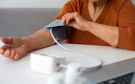 Пожилая женщина измеряет давление тонометром на руке, сидя за столом